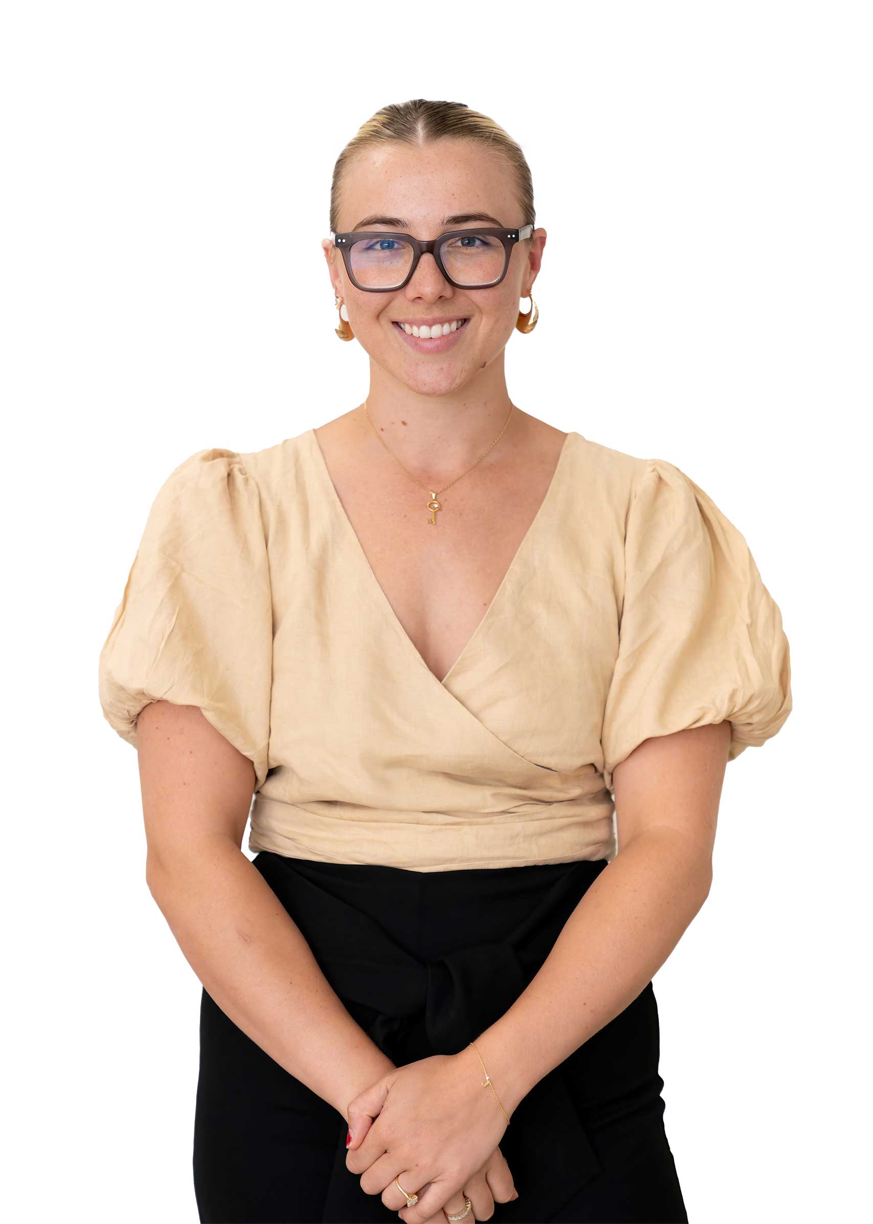 A smiling woman with blonde hair wearing glasses, a beige top, and black trousers, standing against a white background.