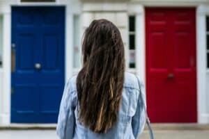 A person with long brown hair in a denim jacket stands facing two doors, one blue and one red.