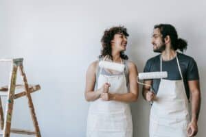 Two people in aprons holding paint rollers smile at each other in front of a white wall; a wooden ladder stands nearby.