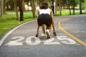 Person in athletic attire poised in a starting position on a road with "2025" painted on it, surrounded by trees.